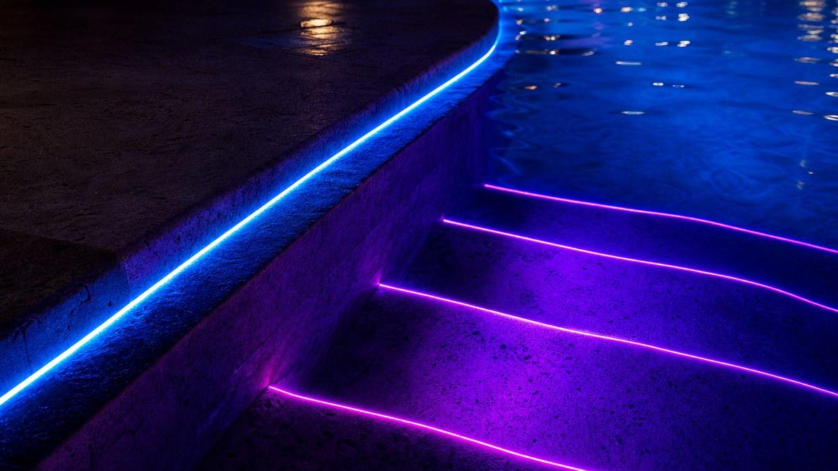 Close-up of side-glow fiber optic pool coping lighting glowing under travertine coping and along lit pool steps in blue and magenta